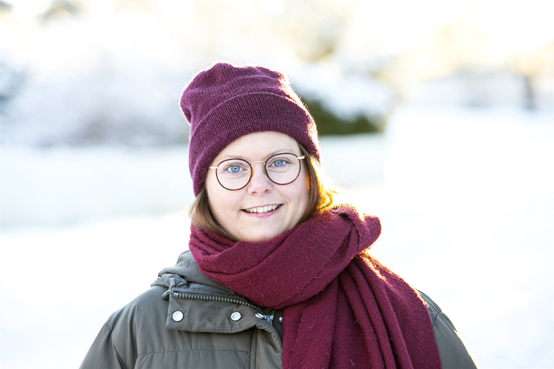 Ann Helen er utendørs og ser på kamera med et smil. Hun har rød lue og rødt skjerf. Uklart vinterlandskap bak.