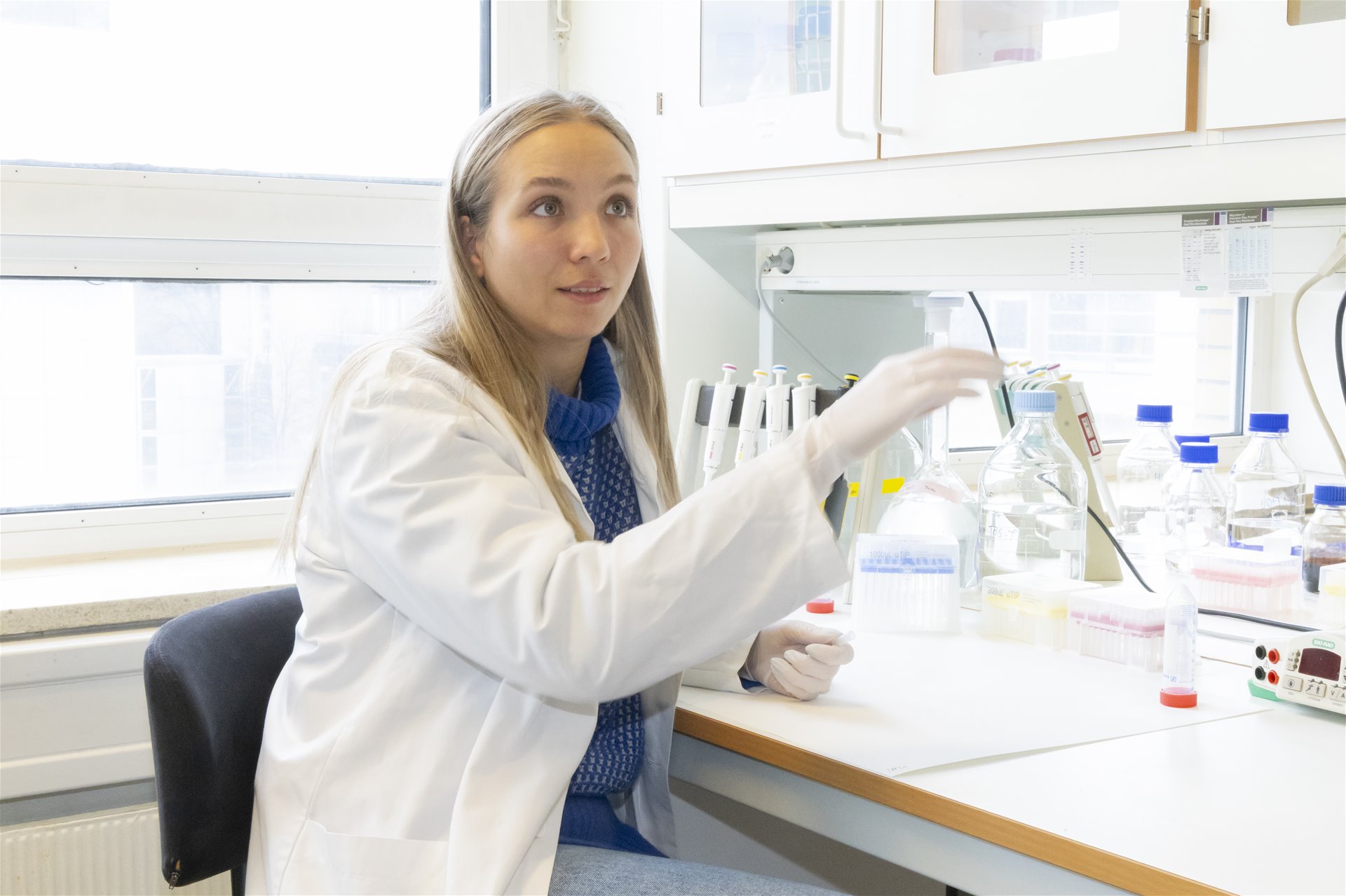 Thea sitter ved bordet i laboratoriet, iført labfrakk. Hun gestikulerer og smiler.