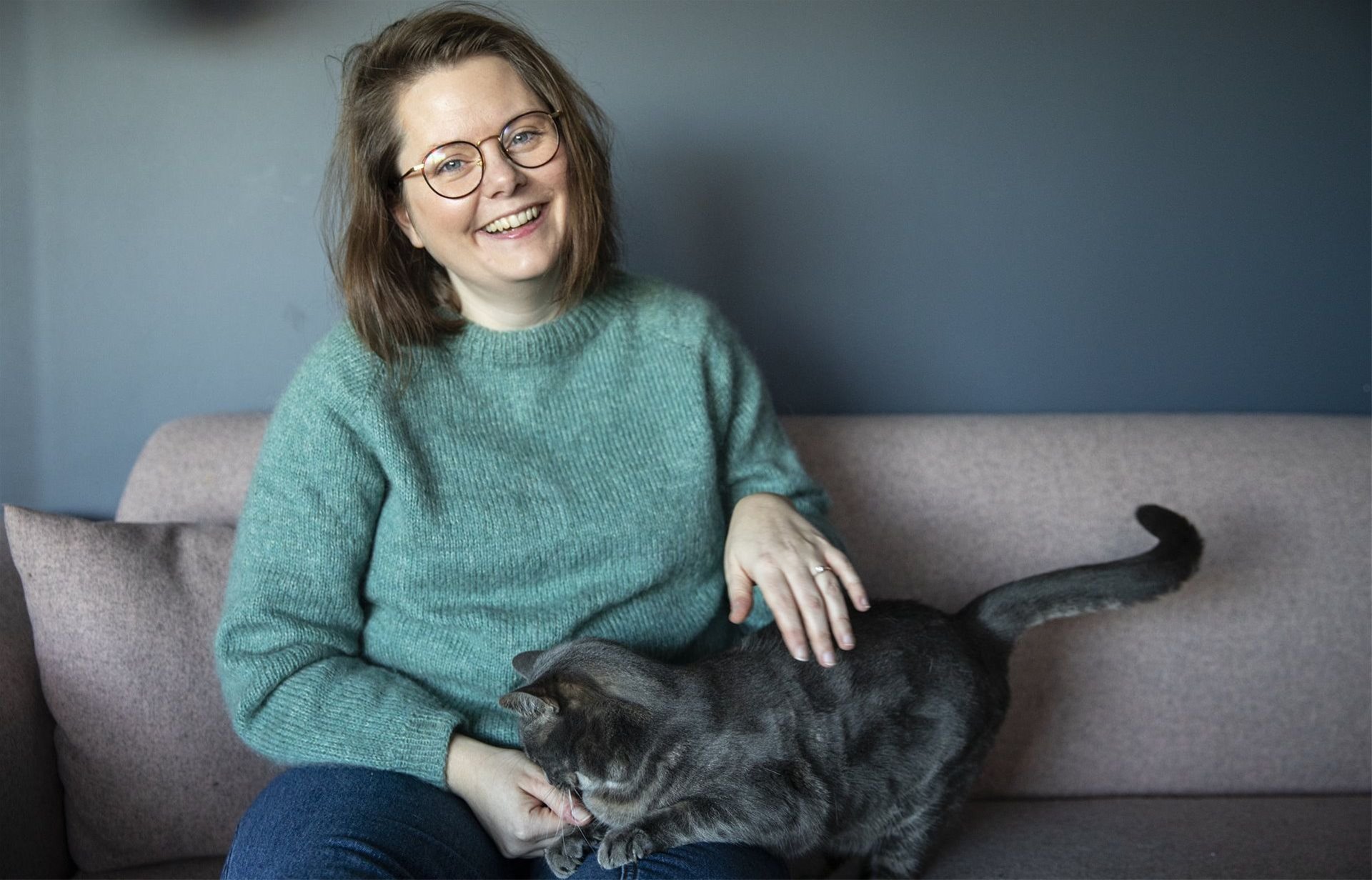 Ann Helen sitter i sofaen med sin grå katt i fanget, hun smiler