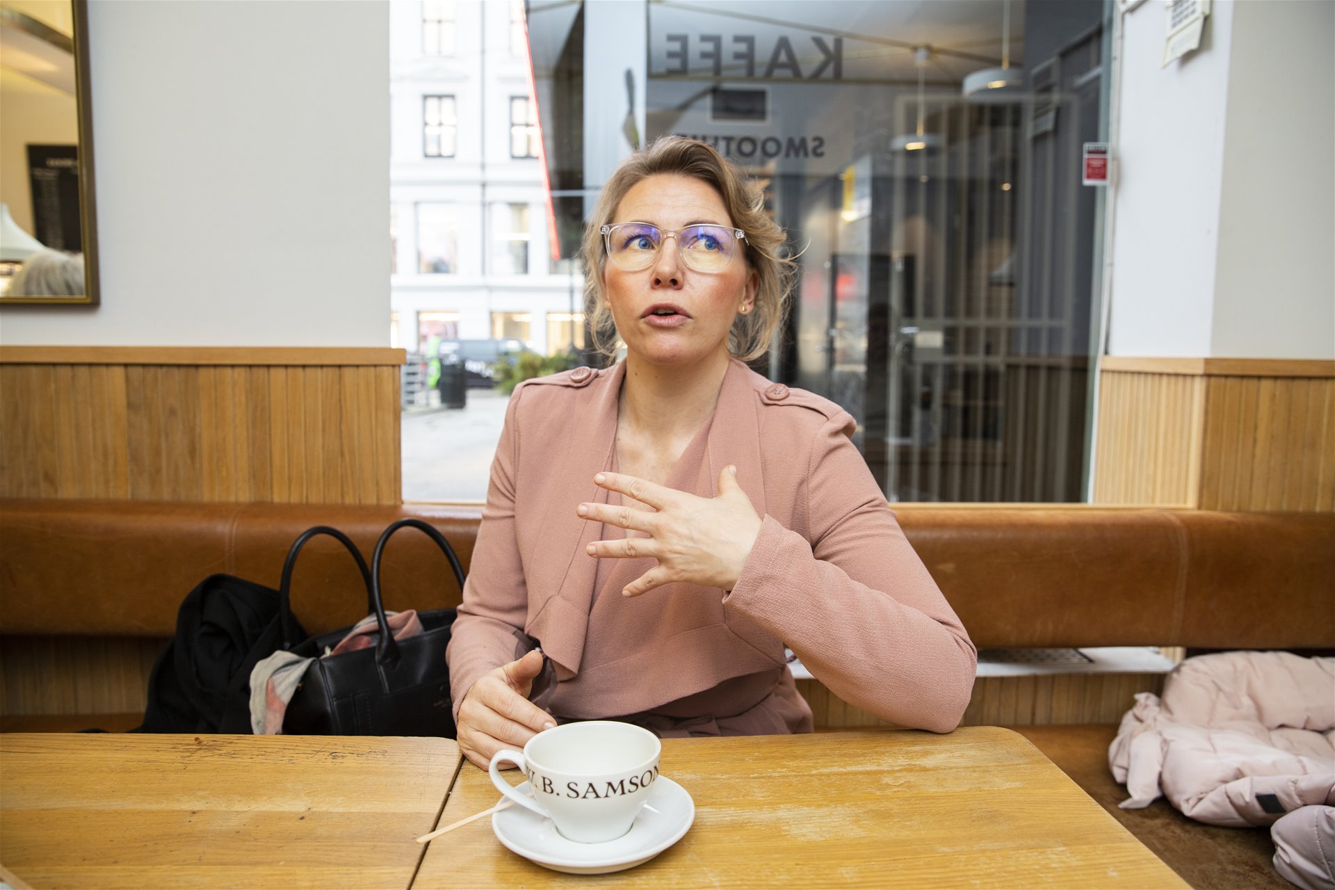 Renate sitter ved bordet og snakker engasjert med armene. Det står en hvit tekopp på bordet.