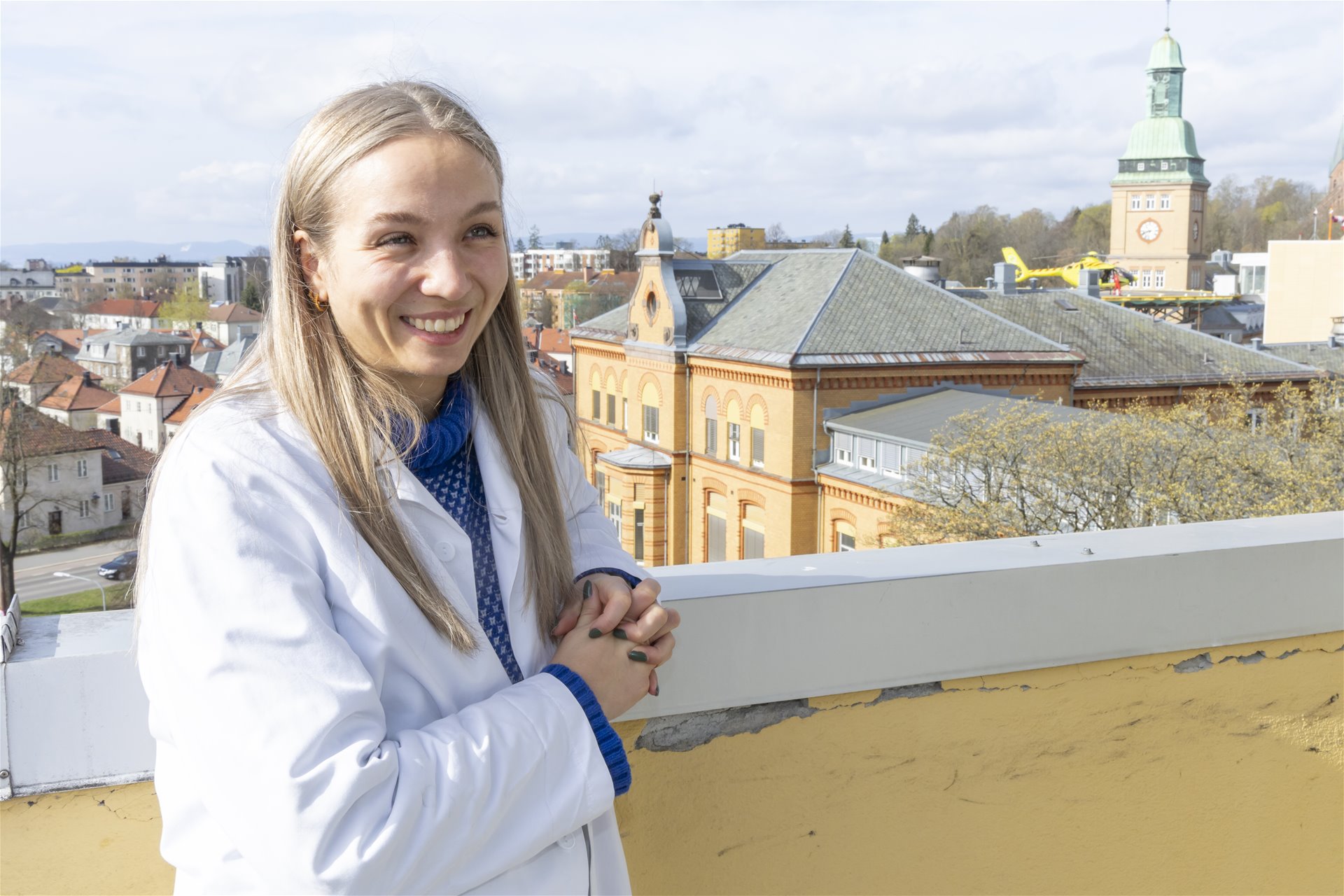 Thea står på taket av Ullevål sykehus og smiler. I bakgrunnen sees tak, helikopterlandingsplassen og klokketårnet.